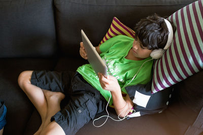 High angle view of boy listening music through digital tablet while reclining on sofa at home