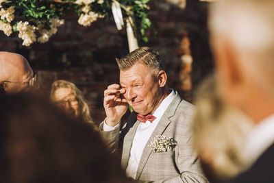 Smiling groom wiping his tears with finger amidst guests on sunny day