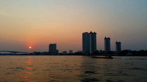 Modern buildings by sea against sky during sunset