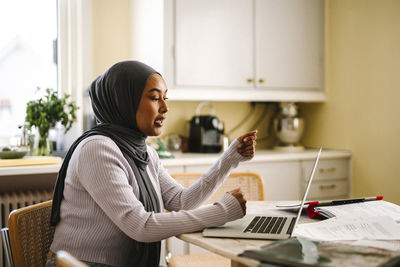 Freelancer wearing hijab doing video call through laptop sitting at table in kitchen