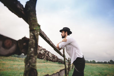 Bearded young man wearing hat standing by fence on field