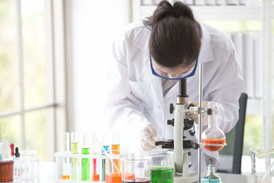 Woman using microscope by test tubes and beaker with liquids on table