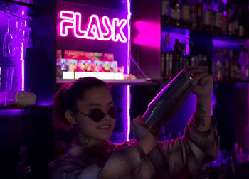 Woman bartender with cocktail shaker in nightclub bar with neon light, barman at club party