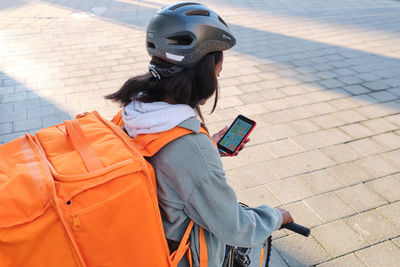 African american female cyclist with thermo backpack standing on paved sidewalk with bicycle while checking location information on cellphone