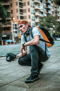 Young man looking away while holding camera on footpath in city