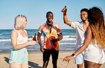 Positive young ethnic guy in casual clothes and sunglasses with beer bottle in hand proposing toast and smiling while spending time on sandy seashore with cheerful multiracial friends