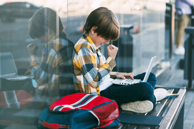 Boy using laptop while sitting on bench against modern building