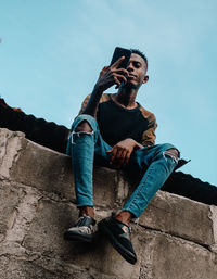 Full length of young man sitting on wall using a mobile phone against sky