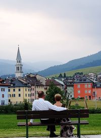 Rear view of people sitting on bench by buildings against sky