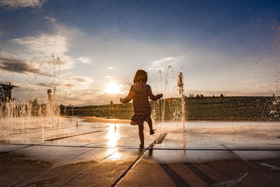 Rear view of woman standing by fountain against sky during sunset