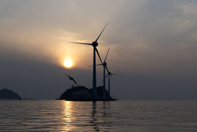 Silhouette of wind turbines in sea against sunset sky