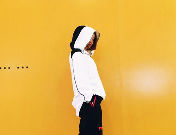 Side view of woman wearing hood while standing against yellow wall