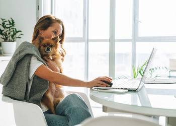 Young adult blonde woman with long hair working on laptop sitting on kitchen with dog at home