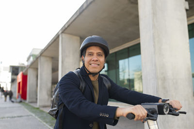 Smiling businessman wearing helmet and riding electric push scooter on sidewalk