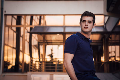 Portrait of young man standing against wall during sunset