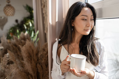 Asian woman drinking coffee while standing near the window in the morning.