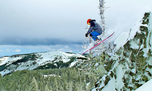 Full length of skier jumping over snowcapped mountains against cloudy sky