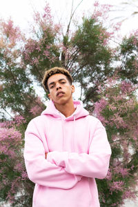 Low angle of confident young ethnic man with dyed afro hairstyle dressed in trendy pink hoodie standing with arms crossed against blooming tree and looking at camera