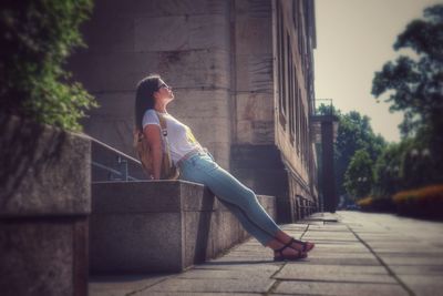 Side view of young woman with backpack sitting on steps in city