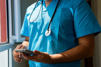 Unrecognizable crop doctor in uniform using mobile phone while standing in corridor near window in hospital