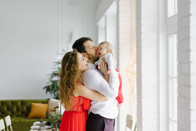 A beautiful family with a baby in their arms stand near the window in the living room, decorated 