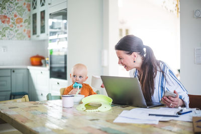 Smiling working mother talking with daughter while freelancing at dining table in kitchen