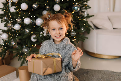 A little girl child in a knitted sweater holds a christmas gift box on holiday in a decorated house