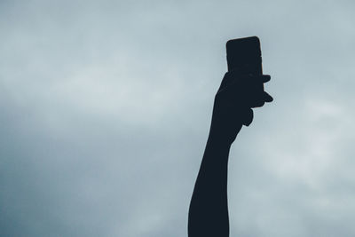 Low angle view of silhouette hand holding phone against cloudy sky