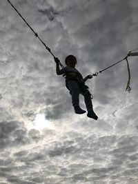 Low angle view of boy zip lining against cloudy sky