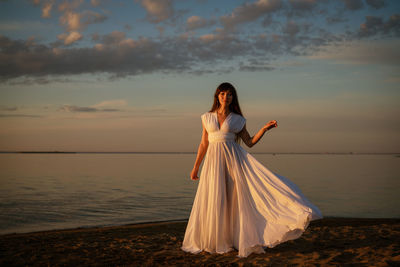 Beautiful young woman in a white long dress near the sea at sunset stands