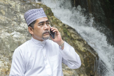 Thoughtful man talking on phone while sitting against waterfall