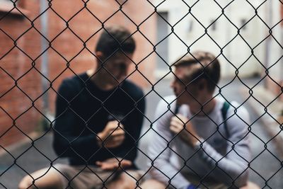 Male friends talking while sitting outdoors seen through chainlink fence