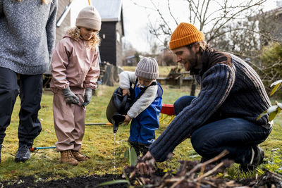 Man guiding son using watering can by girl while doing gardening at back yard