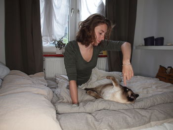 Woman sitting by siamese cat on bed at home