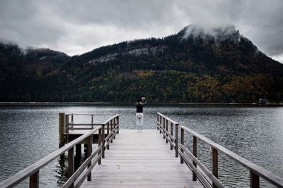 Rear view of woman standing on pier over lake against cloudy sky