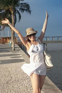Full length of a young woman, smiling, with arms up in the air over a tropical beach scenery 