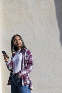 Portrait of smiling young woman using phone while standing against wall