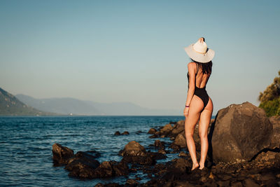 Rear view of sensuous woman standing on rocks at beach against clear sky