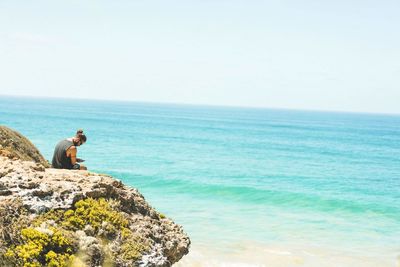 Side view of man sitting on cliff by sea against sky during summer