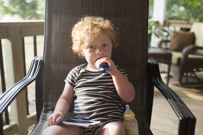 Portrait of cute baby boy eating food while sitting with smart phone and milk bottle on chair at porch