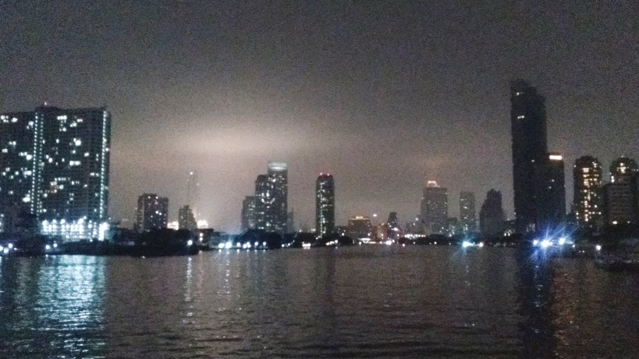 ILLUMINATED BUILDINGS BY RIVER AGAINST SKY AT NIGHT