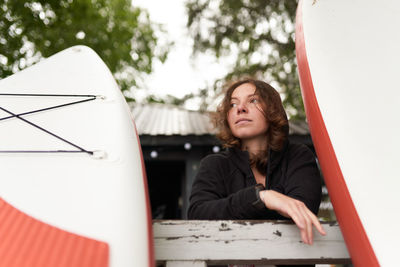 Low angle of calm female surfer stranding on wooden embankment with paddle board while relaxing after training and looking away
