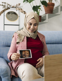 Smiling pregnant woman showing ultrasound on video call over laptop at home