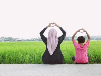 Rear view of mother with daughter making heart shape with hands by grassy field