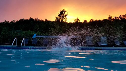 Swimming pool by trees against sky during sunset