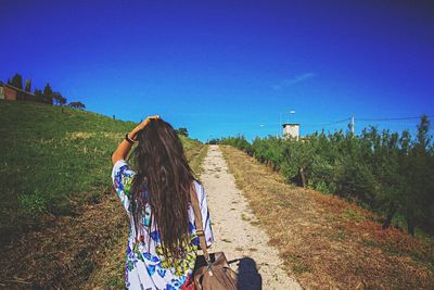 Rear view of woman standing on trail amidst plants against clear blue sky