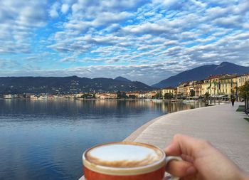 Cropped image of man holding coffee cup at pier by lake garda against cloudy sky