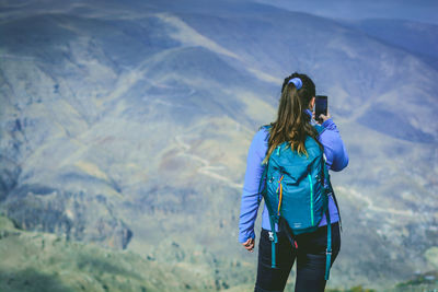 Rear view of woman photographing with mobile phone while standing against mountains