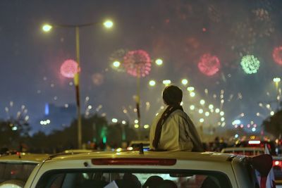 Rear view of boy looking at firework display while sitting on car roof at night