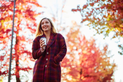 Young woman holding coffee while standing against autumn trees